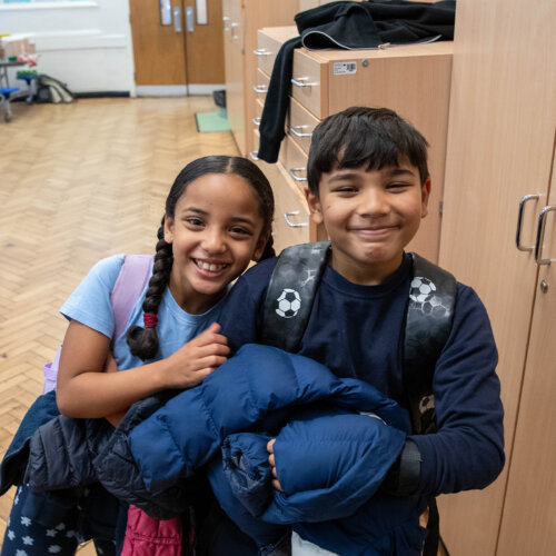 A photo of two childen smiling at the camera at school. The background is a table full of breakfast cereal.